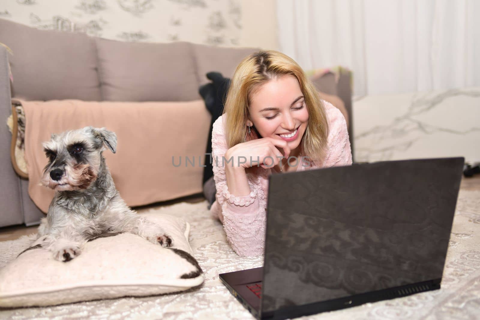 Young blonde woman is laughing at home with dog while chatting with someone through a video call on a laptop.