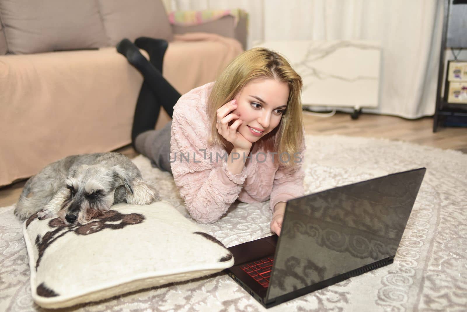 Lovely woman in a pink sweater is lying on the carpet with a laptop at home, a gray dog is sleeping on a pillow next to her. by Nickstock