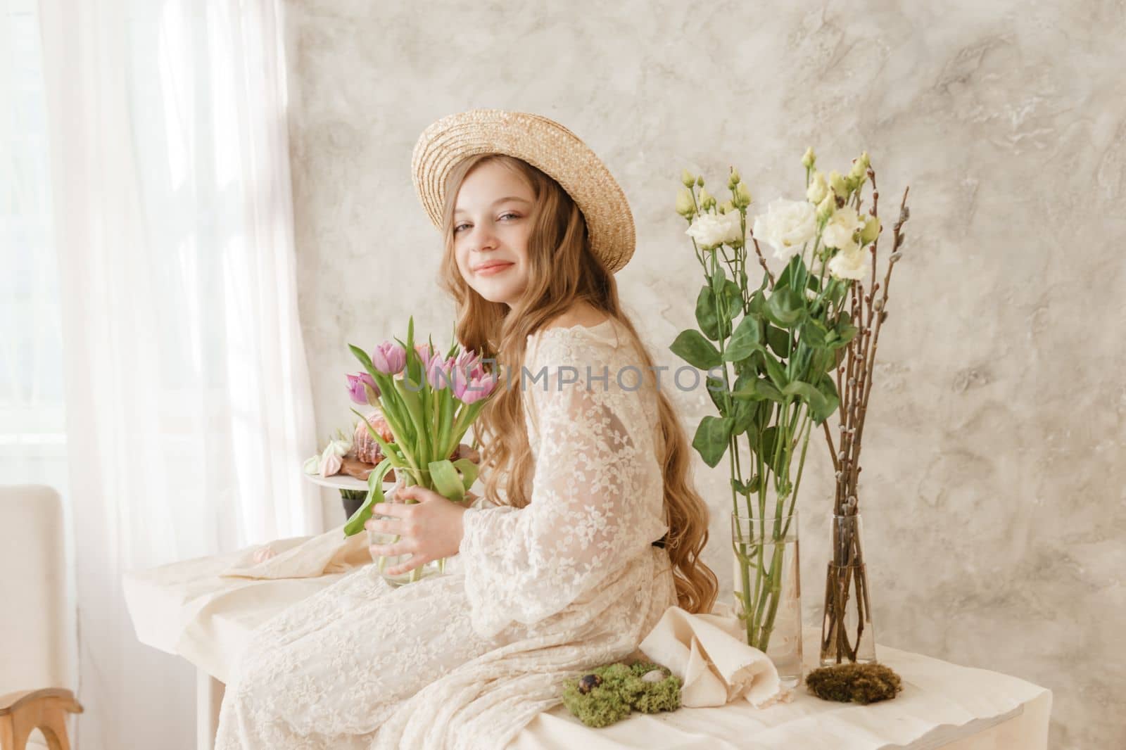 A girl with long hair in a dress and hat is sitting at the Easter table with cakes, spring flowers and quail eggs. Happy Easter celebration
