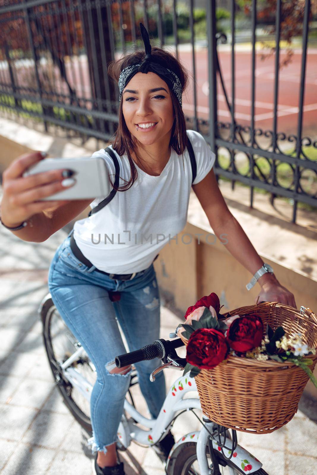 Smiling young woman taking selfie, riding the bike with flower basket, on a sunny day.