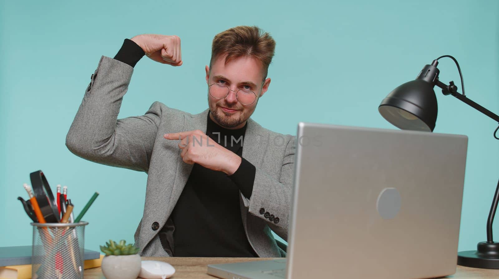 I am strong and independent. Happy business man sitting at office desk showing biceps and looking confident, feeling power strength, energy to gain success win. Young satisfied guy on blue background