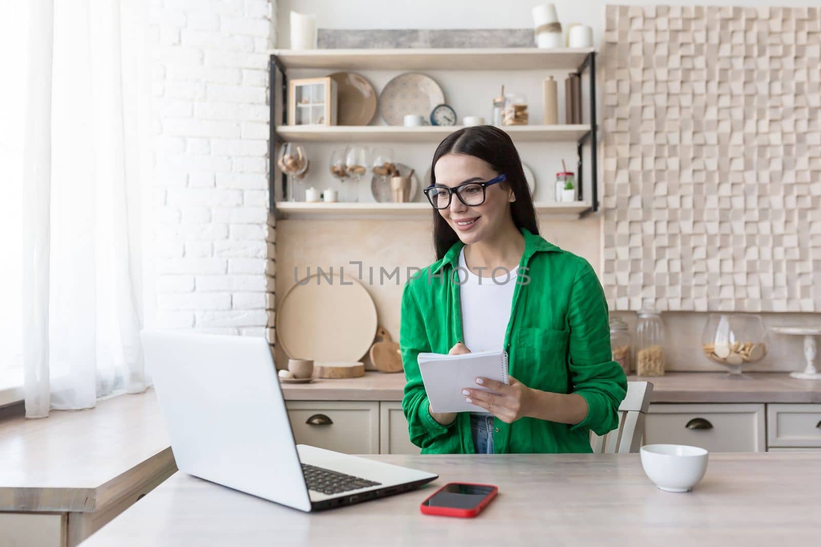 A young beautiful woman is having a remote job interview online. Sitting at home in the kitchen with a laptop. Speaks through a video call, introduces himself, passes the exam, writes in a notebook.