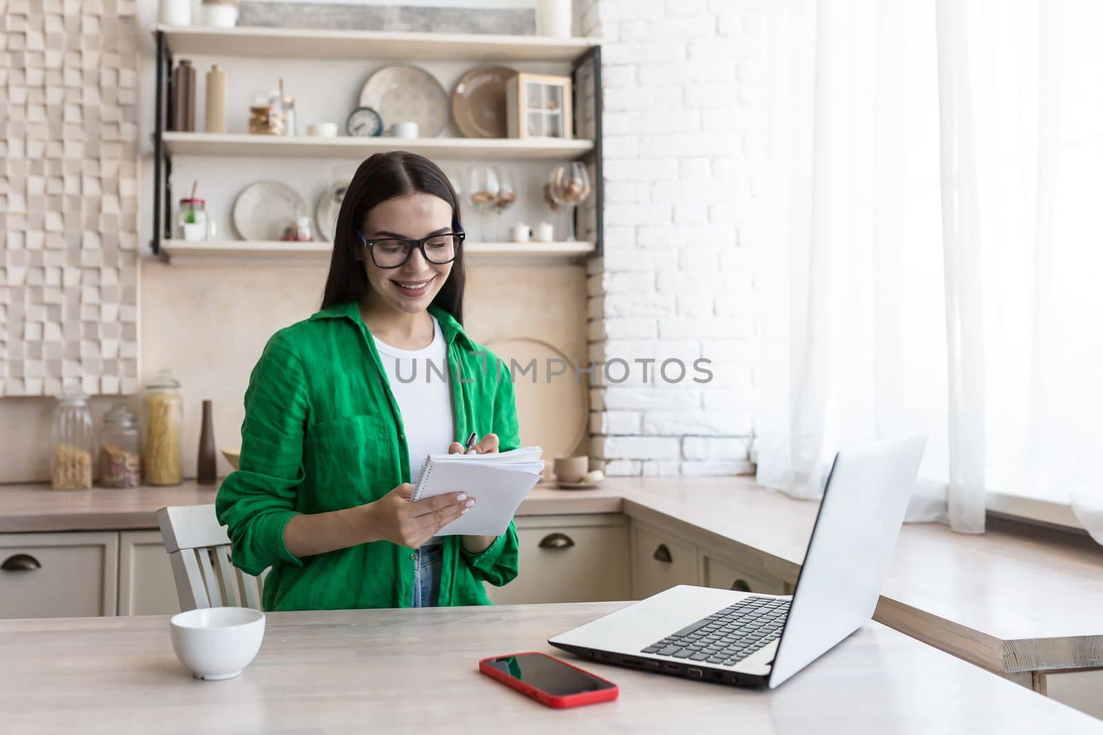A young female journalist, writer, blogger works remotely from home using a laptop online. Sitting in the kitchen at the table, making notes in a notebook.
