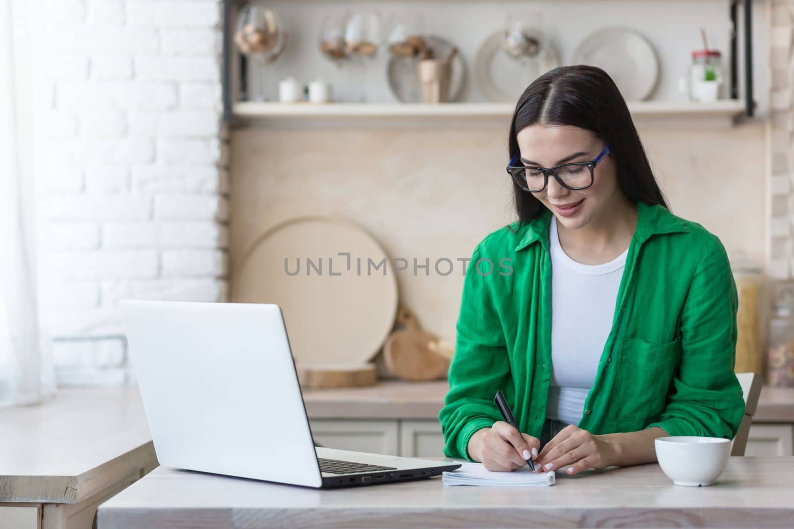 A young woman journalist, writer, blogger works remotely from home using a laptop online. Sitting in the kitchen at the table, making notes in a notebook.