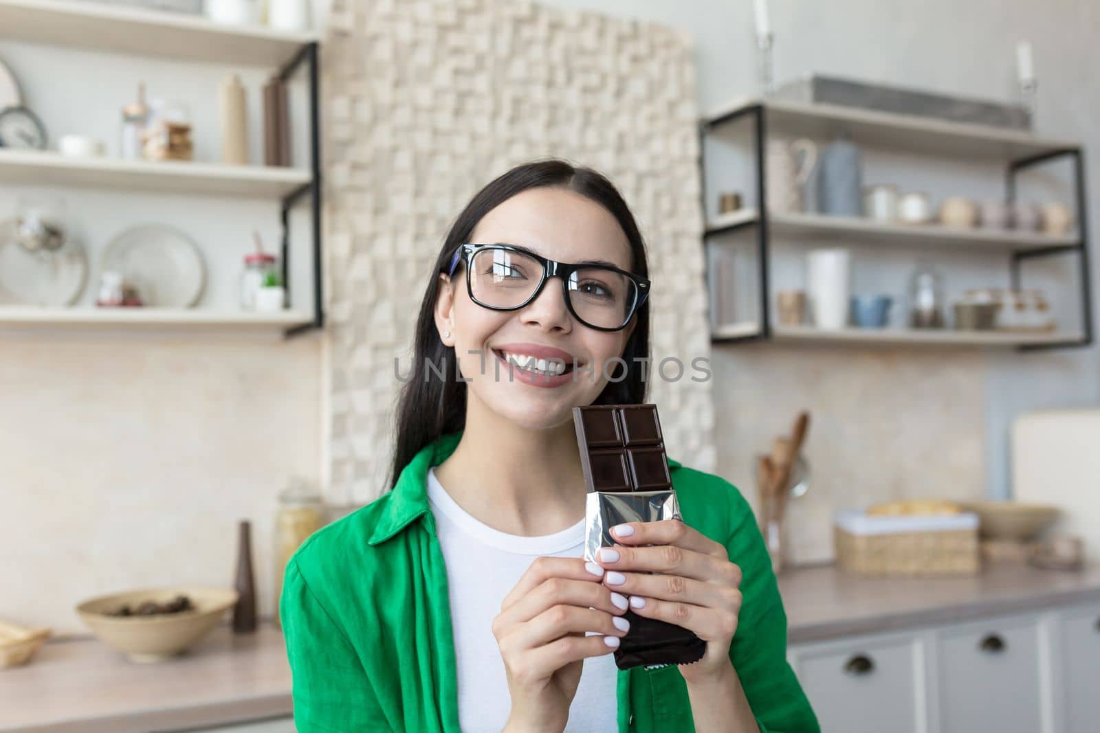 Close up photo. Portrait of a young beautiful woman holding a bar of black chocolate in her hands. Standing at home in the kitchen, looking at the camera, smiling.