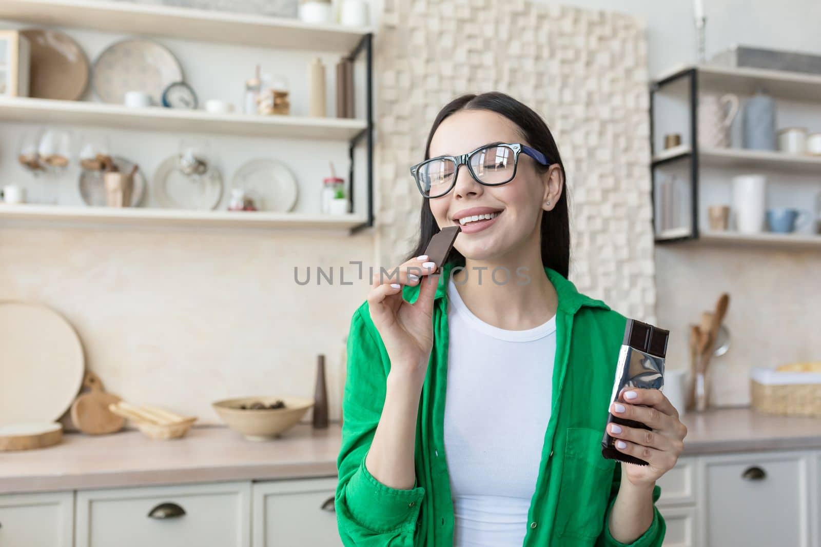 Portrait of a young beautiful woman in glasses eating a piece of black chocolate, holding a tile in her hands. Standing at home in the kitchen, eyes closed, enjoying.
