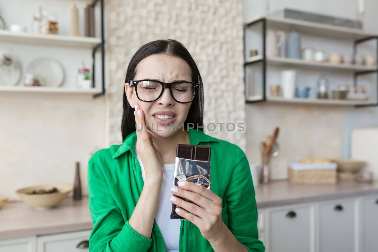 A young woman is sitting in the kitchen at the table at home, eating a bar of dark chocolate. Holds his cheek, feels a strong toothache from salty. She grimaced in pain.