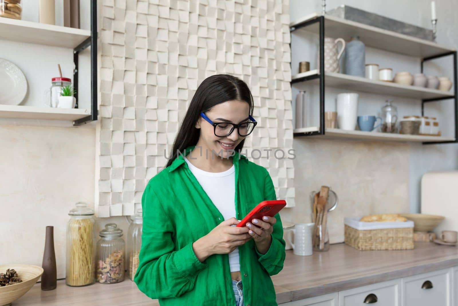 Happy and smiling young teenage girl standing at home in the kitchen and using the phone. Receives messages, chats with friends, boyfriend, chats news, blogs, social networks, online shopping.