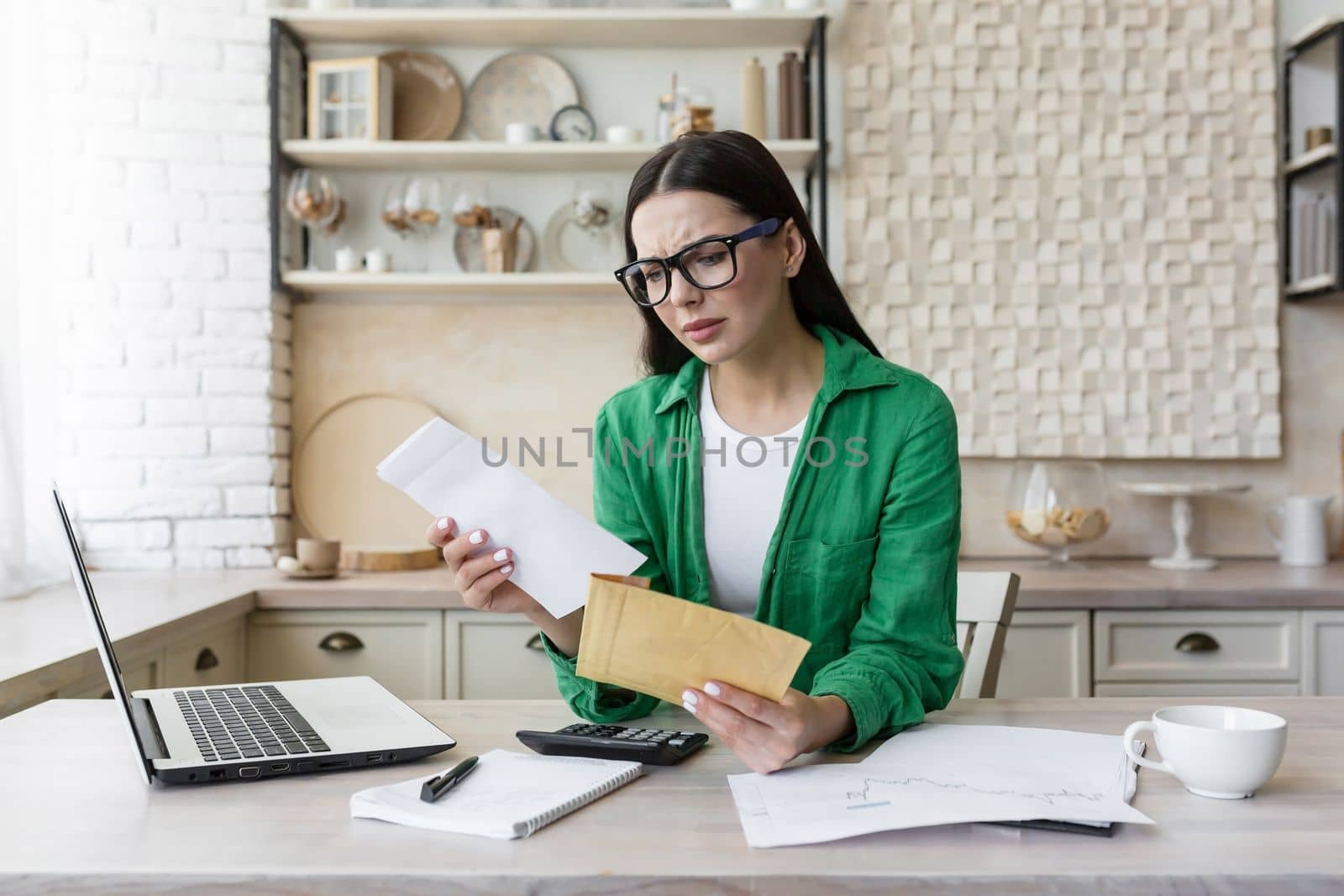 Worried young woman working in the kitchen at home with a laptop. She is holding an envelope with a letter in her hands, she received bad news, an account, debts, divorce papers.