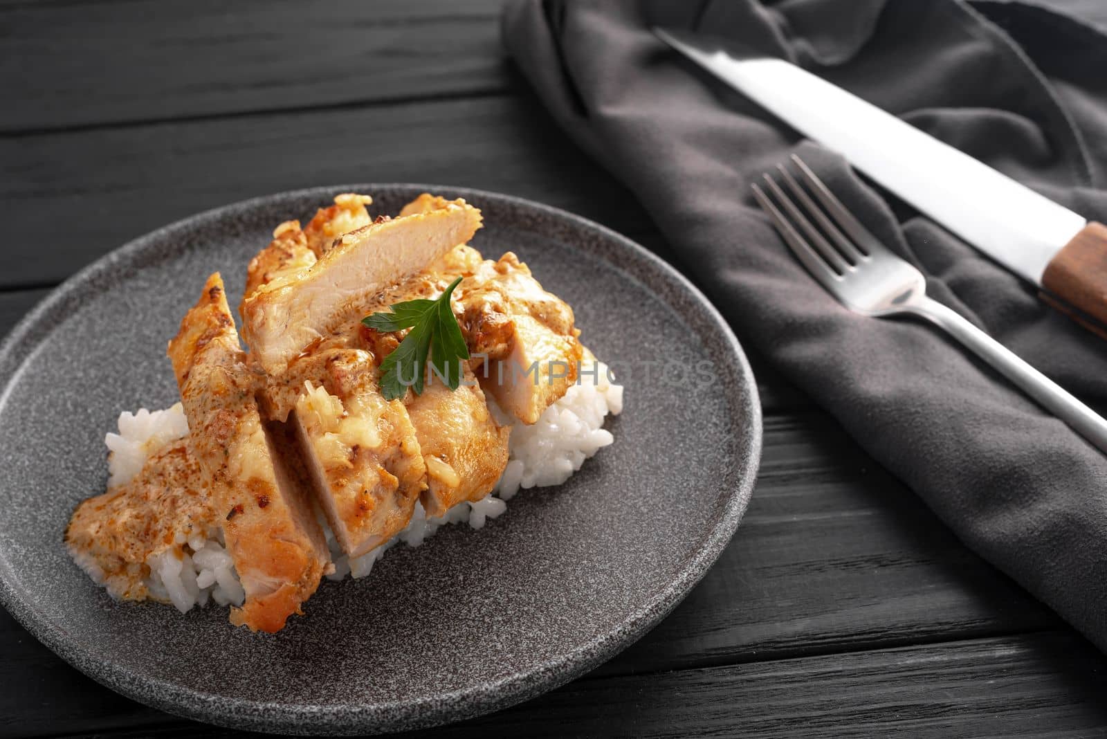 In a creamy sauce, a chicken breast with rice sliced into pieces on a dark background in a gray plate. Next to it is a knife and fork