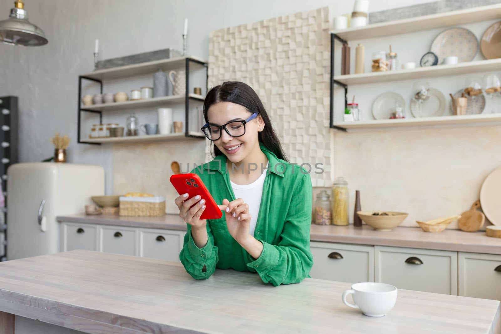 Smiling young woman sitting in the kitchen at home and using the phone. Drinks coffee, eats breakfast, types messages, chats with friends, working, reads news, blogs, social networks, search engine.