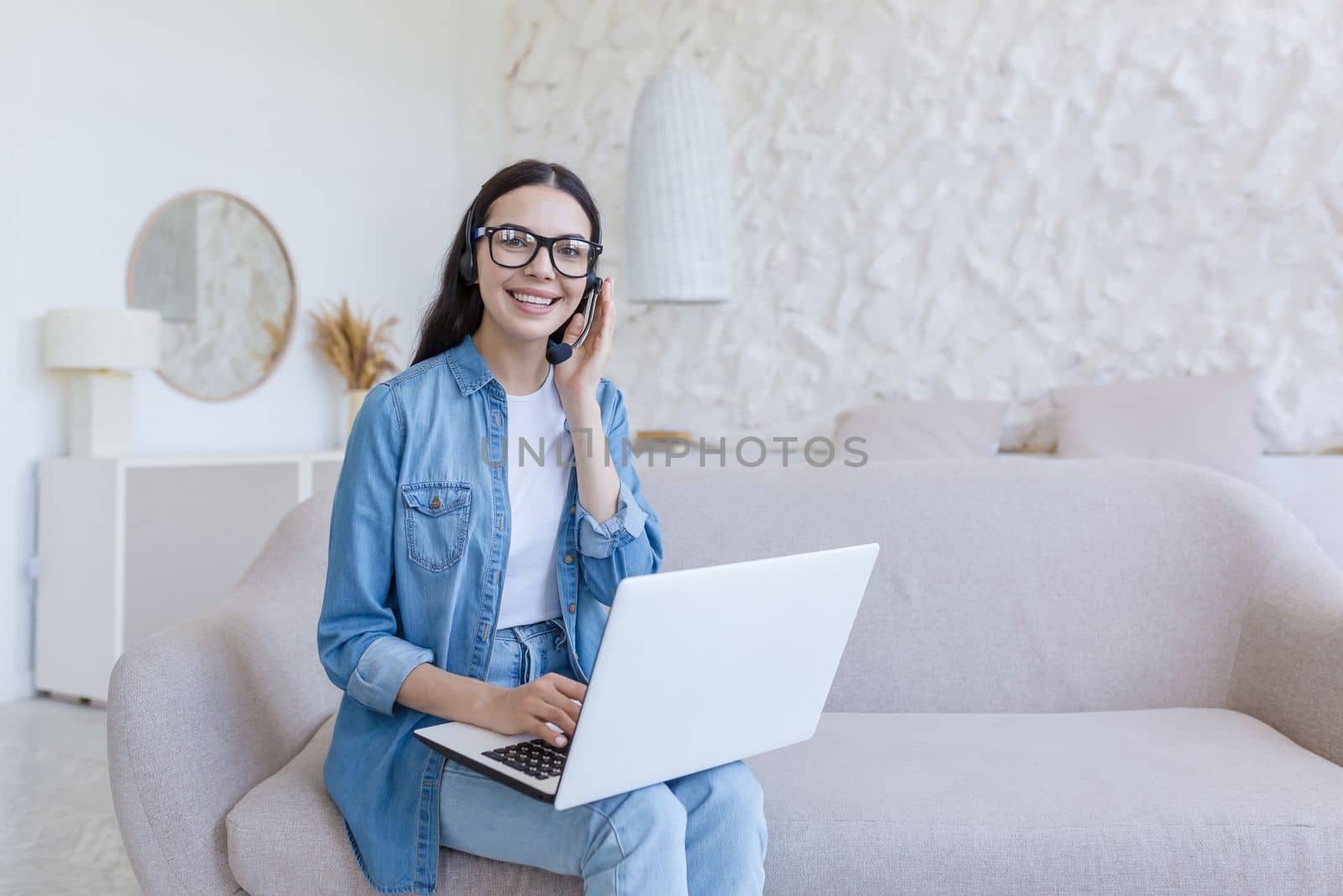 Portrait of a beautiful young female student sitting on the sofa at home with a laptop and a headset. He studies online, remotely. He looks at the camera, smiles.