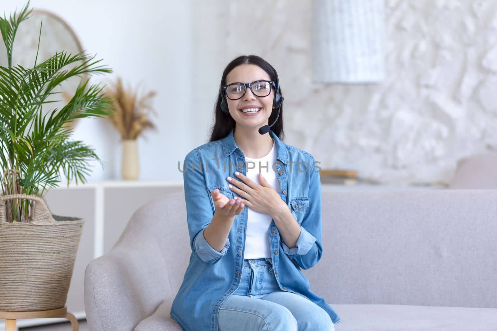 A young woman is having a remote job interview online. Sitting at home on the sofa in a headset. Talks through a video call, introduces himself, looks into the camera, smiles.