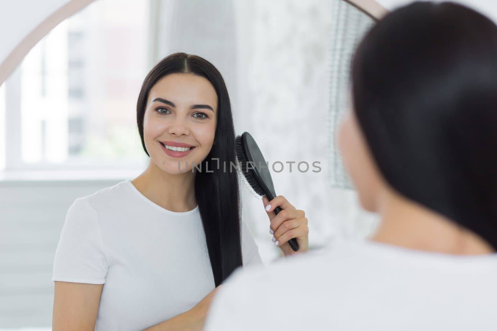 Close-up photo. A beautiful young woman stands and looks at herself in the mirror. She combs her hair with a brush, does her hair. She smiles, looking at the camera.