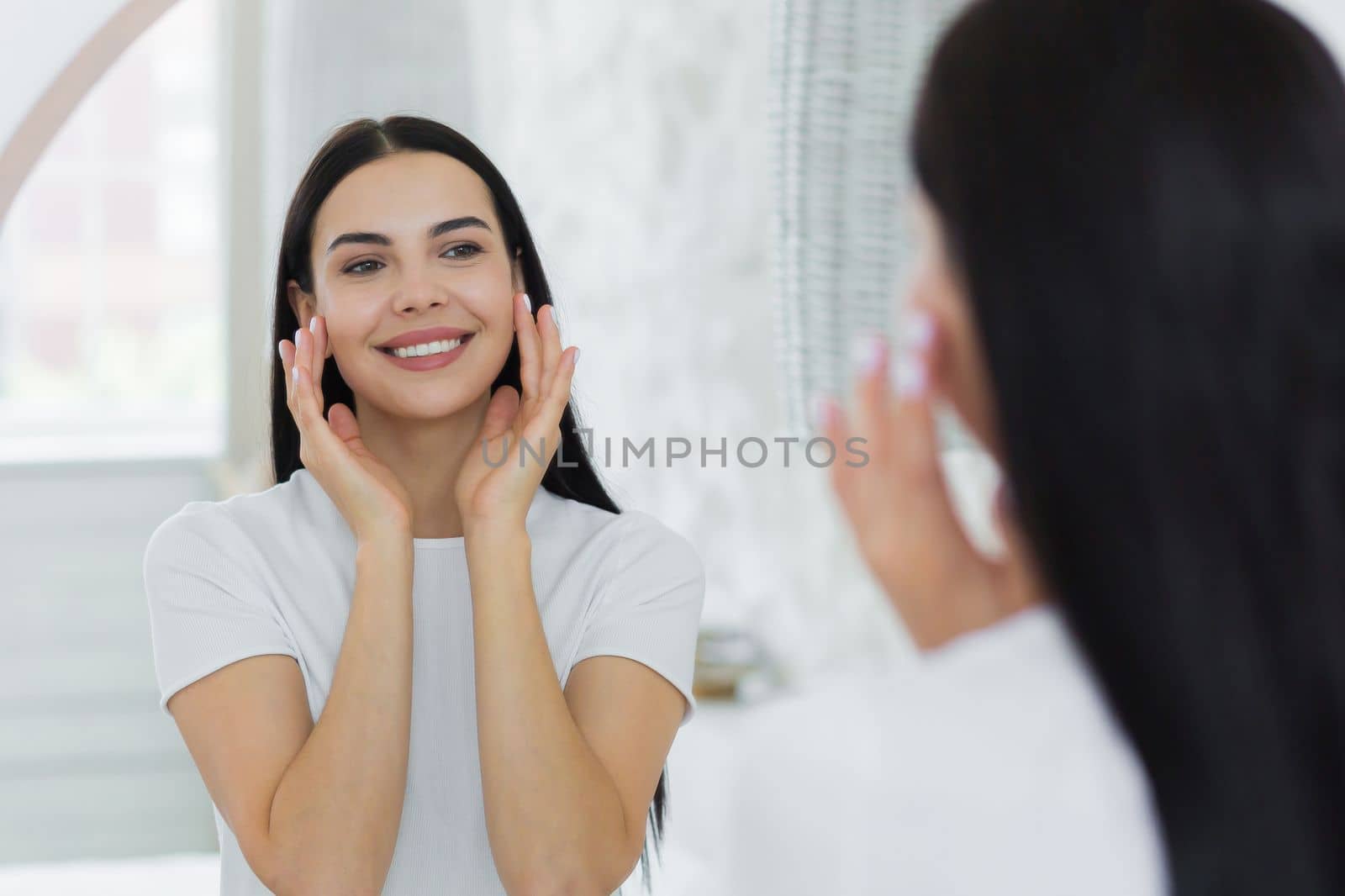 A beautiful young woman stands and looks at herself in the mirror. She touches her face with her hands, applies cream, cosmetics, does a massage, admires herself, smiles.