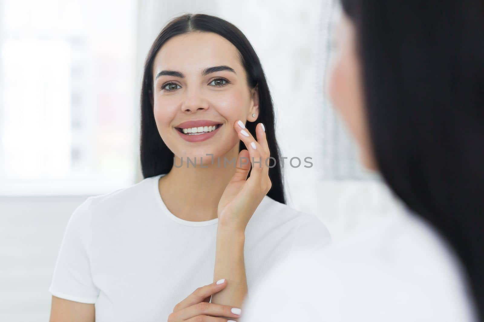 Portrait of a young woman stands and looks at herself in the mirror. She touches her face with her hands, looking at the camera, smilling,