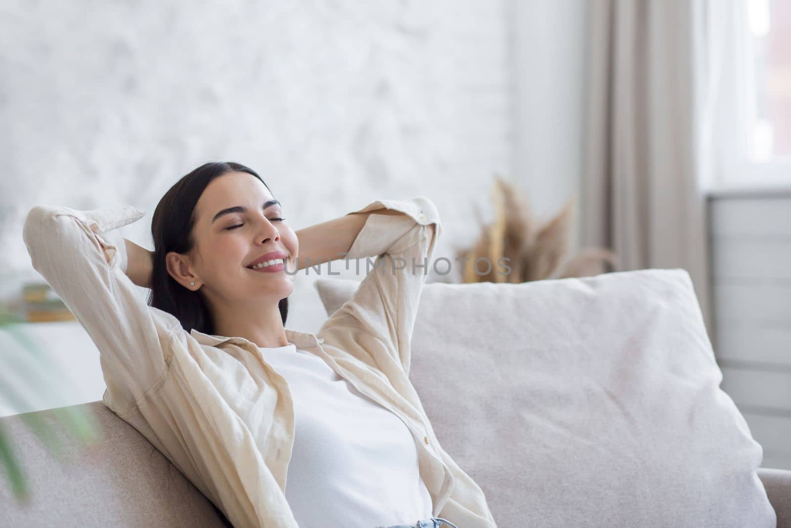 Young beautiful woman resting on sofa at home. She put her hands behind her head, closed her eyes.