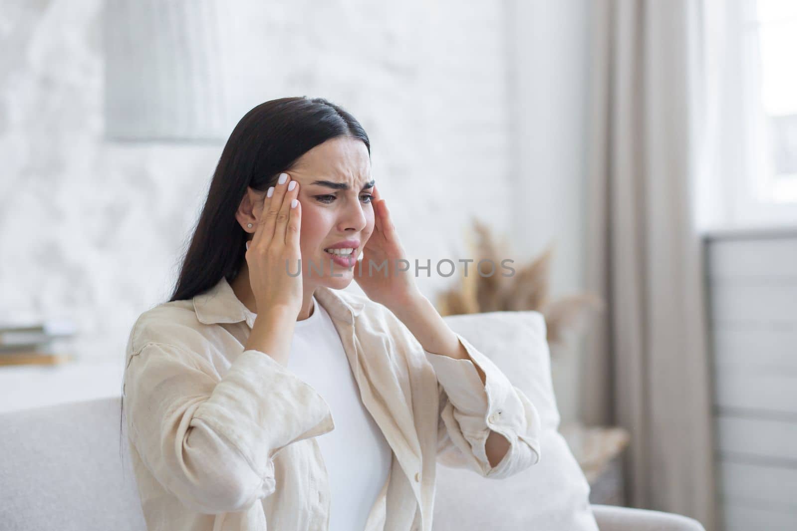 Worried young woman suffering from depression, mental disorders. He is sitting in the hospital room on the sofa near the window, holding his head with his hands, feeling a strong headache, pressure.