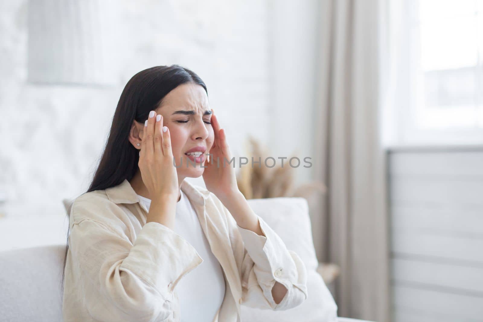A young woman suffers from a severe headache, pressure. She is sitting on the couch at home, holding her head with her hands , doing a massage, wincing in pain.