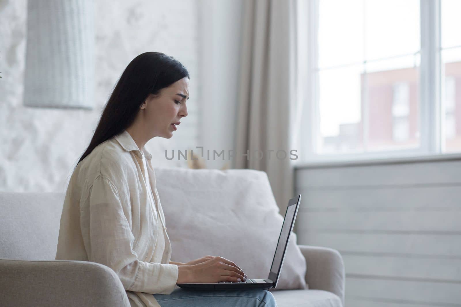 Young upset woman received bad news, documents, divorce letter through online mail. She sits sad at home on the sofa, holds a laptop, cries.