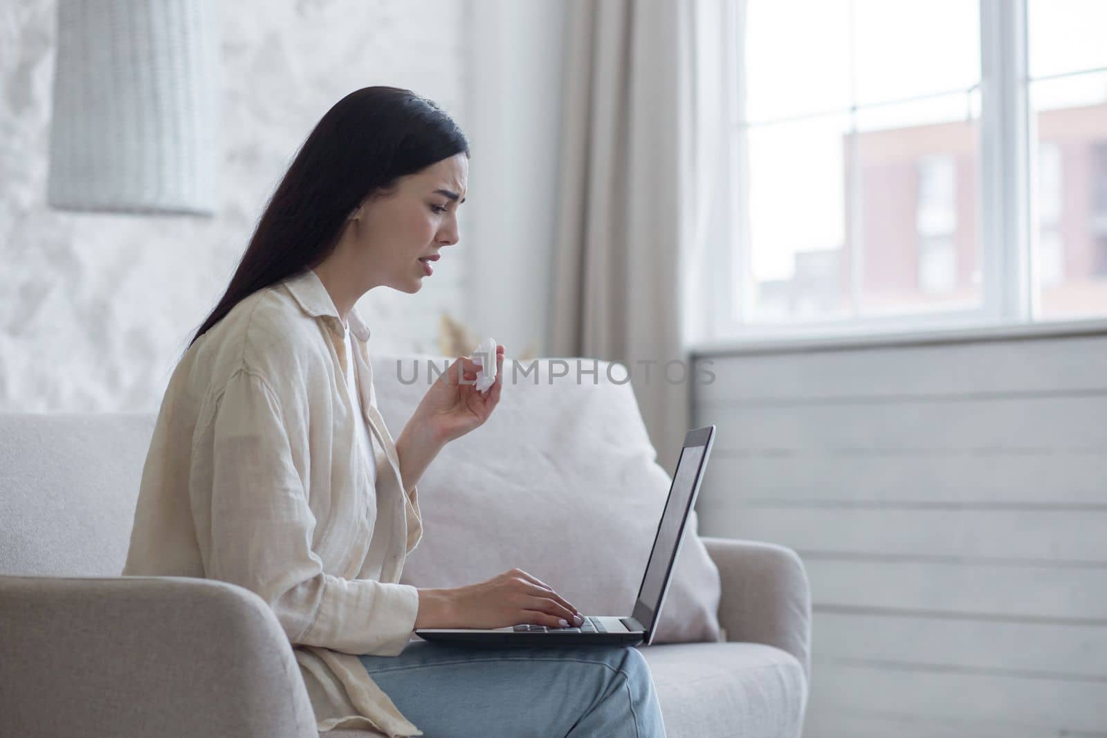 Upset young woman sits at home on the couch by the window with a laptop, reads bad news, received an email. Crying, wiping tears with a napkin.