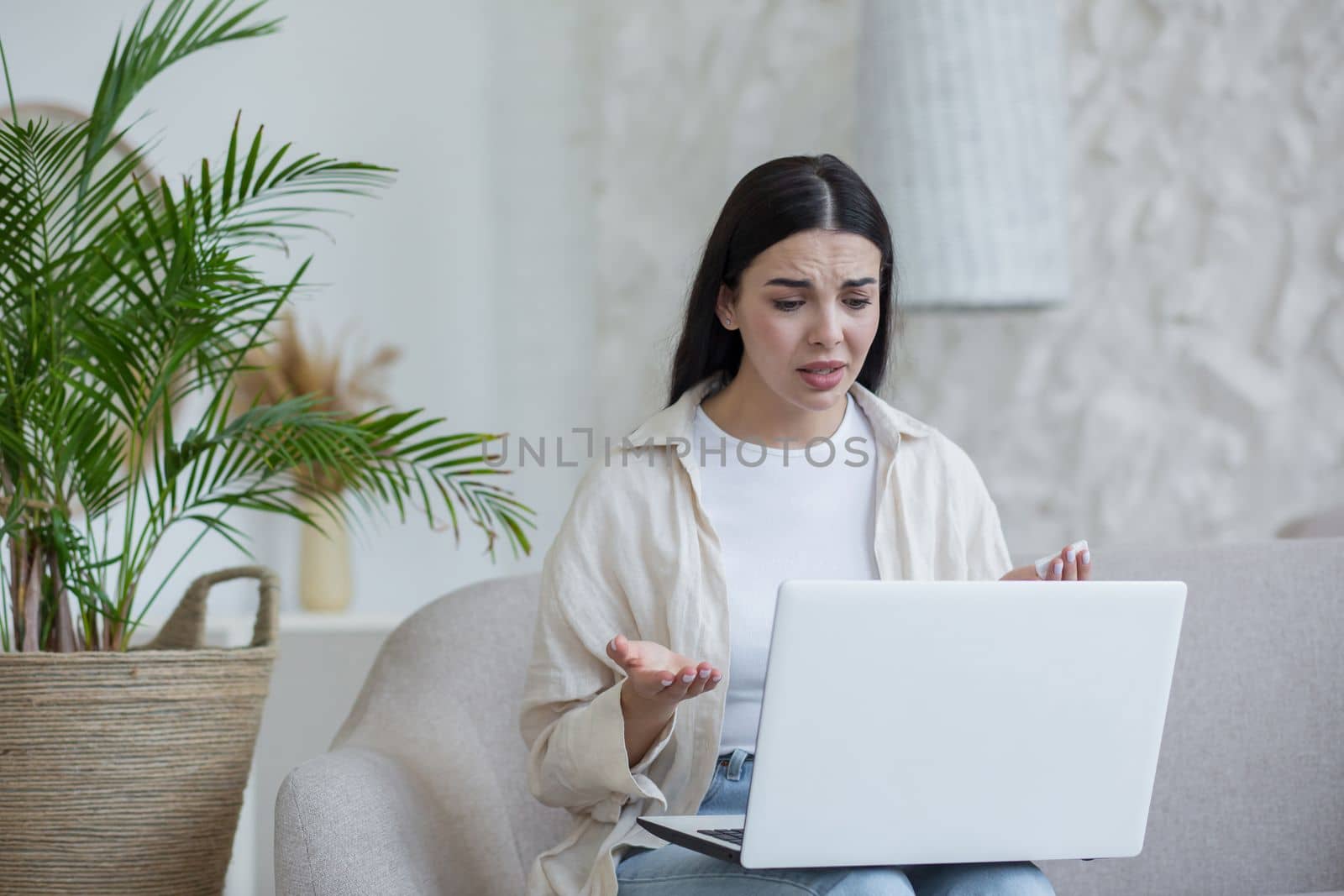 Online psychotherapy. A young worried woman is sitting on the couch at home and holding a laptop on her lap. Consults with a psychologist via video call, solves problems