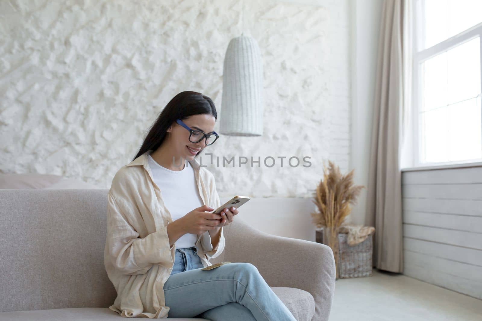 A young beautiful woman in glasses sits on a sofa by the window and uses a mobile phone, dials, texts, reads the news, smiles.
