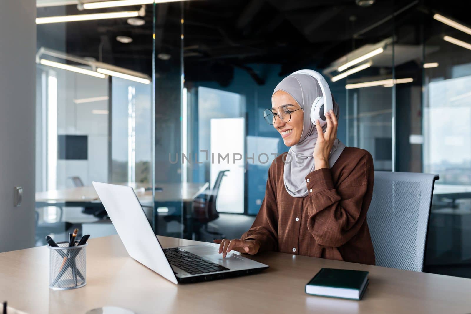 Successful modern businesswoman working inside office with laptop, Muslim woman wearing hijab and headphones listening to audio books and podcasts at workplace, satisfied and successful woman.