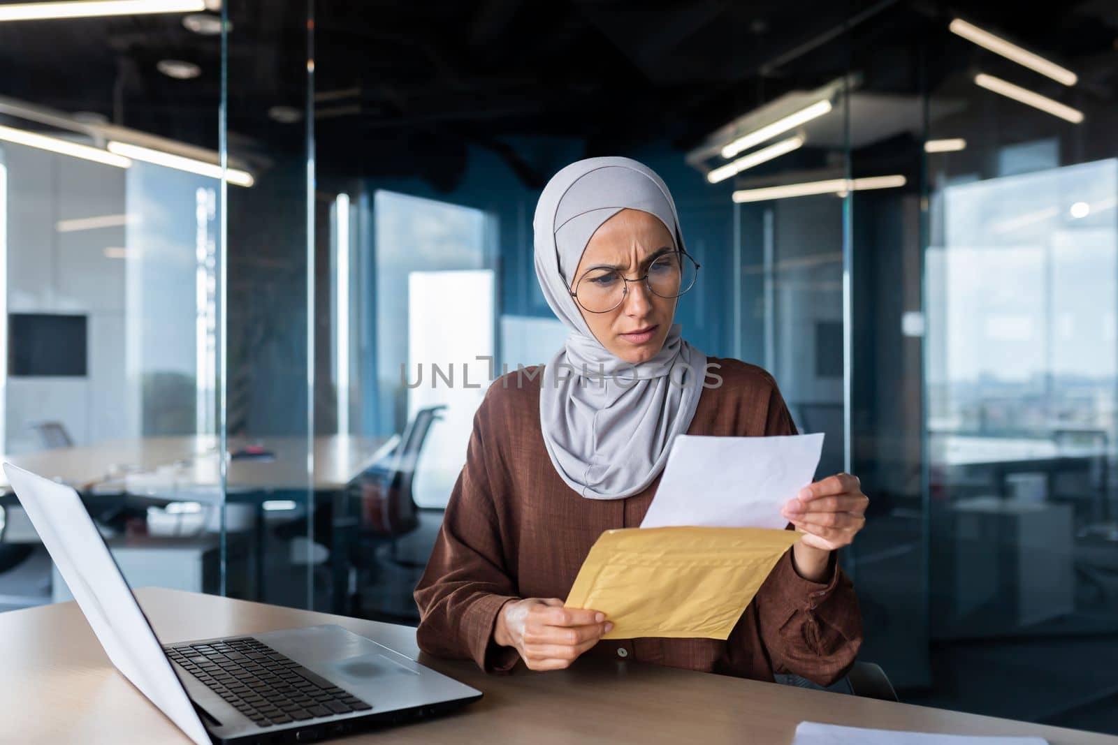 Upset business woman in hijab working inside office, woman received letter envelope mail with bad news, office worker upset by notification working at workplace with laptop.