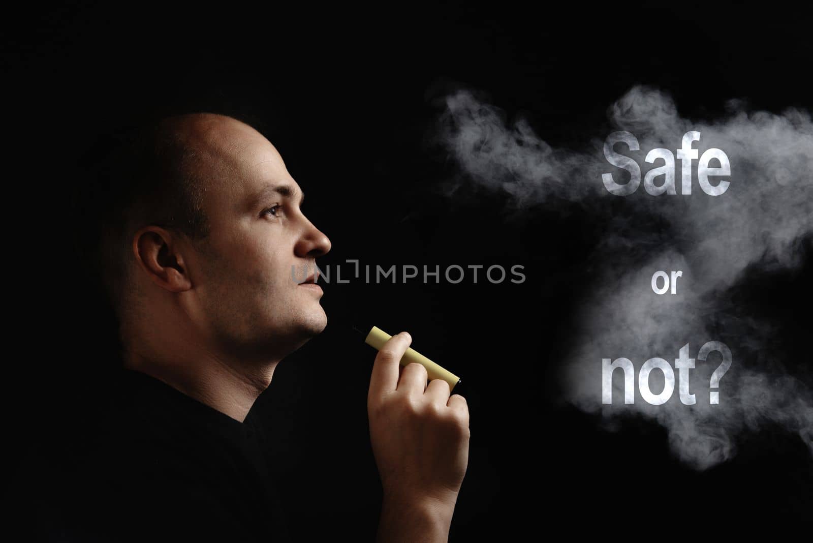 E-cigarette safe or not. A young man smokes an electronic cigarette against a black background and blows smoke. synthetic nicotine