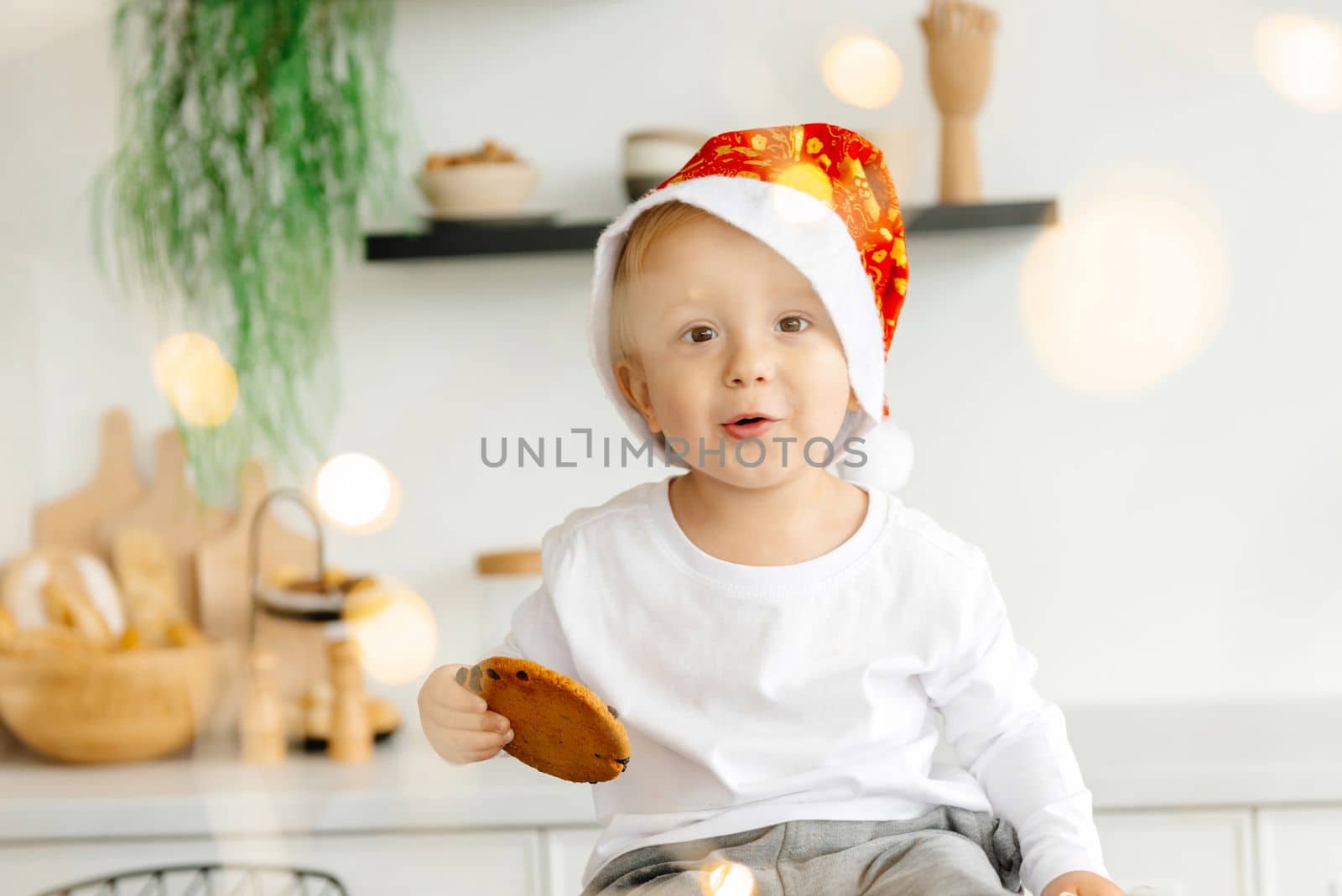 Kid looks at the camera wearing a Christmas hat and holding a cookie .