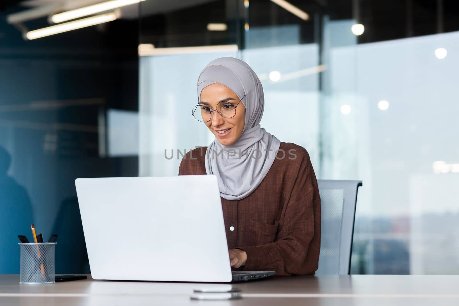 Smiling and dreamy businesswoman working inside office with laptop, woman in hijab and glasses office worker happy and satisfied with work sitting at desk by voronaman