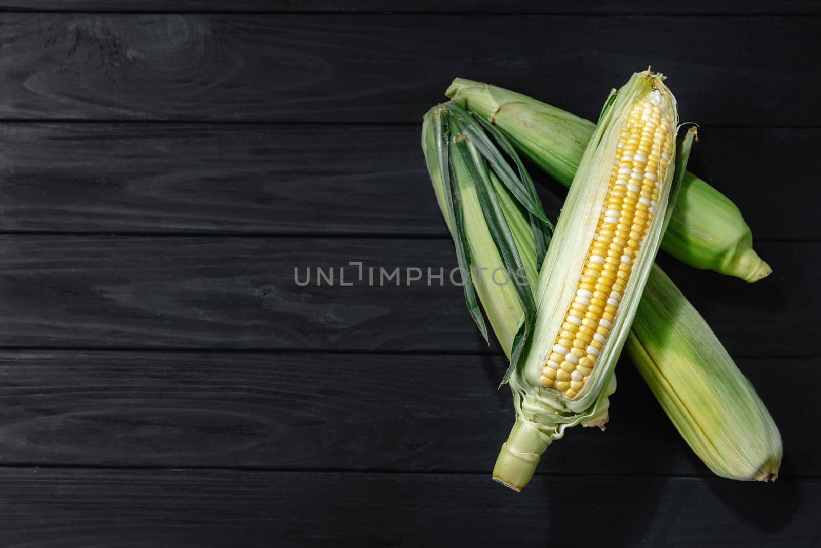 A few corn in leaves lie on a wooden dark background. View from above, space for text.
