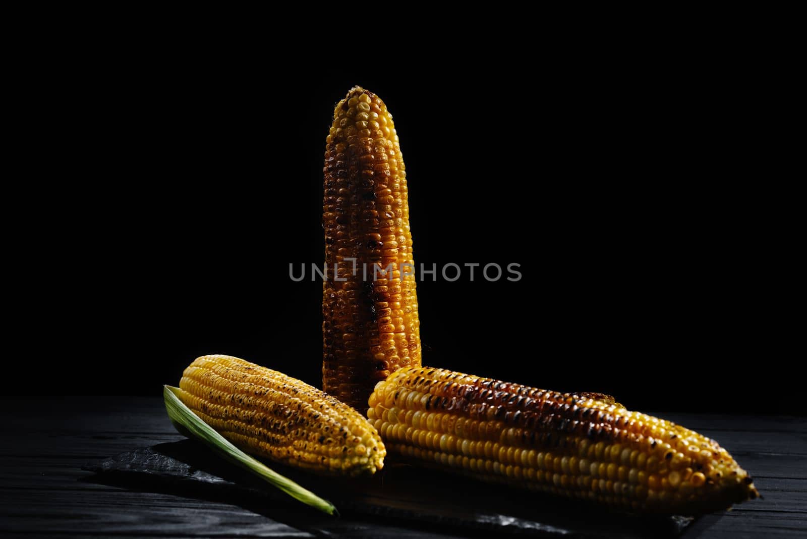 A cob of corn roasted over charcoal on a wooden board. Whole corn cobs. Dark background
