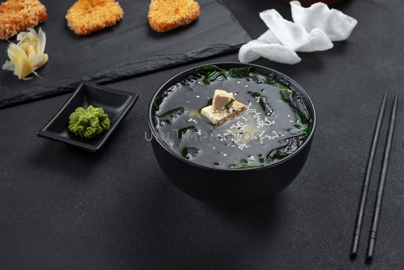 Asian food. Soup on a black background in a black bowl. Soup with rice chips. Top view. Miso soup with tofu and salmon with onions and sesame seeds. Next to it on a slate, chicken nuggets, Chinese chicken. Set or set of Chinese Asian food on a black background.