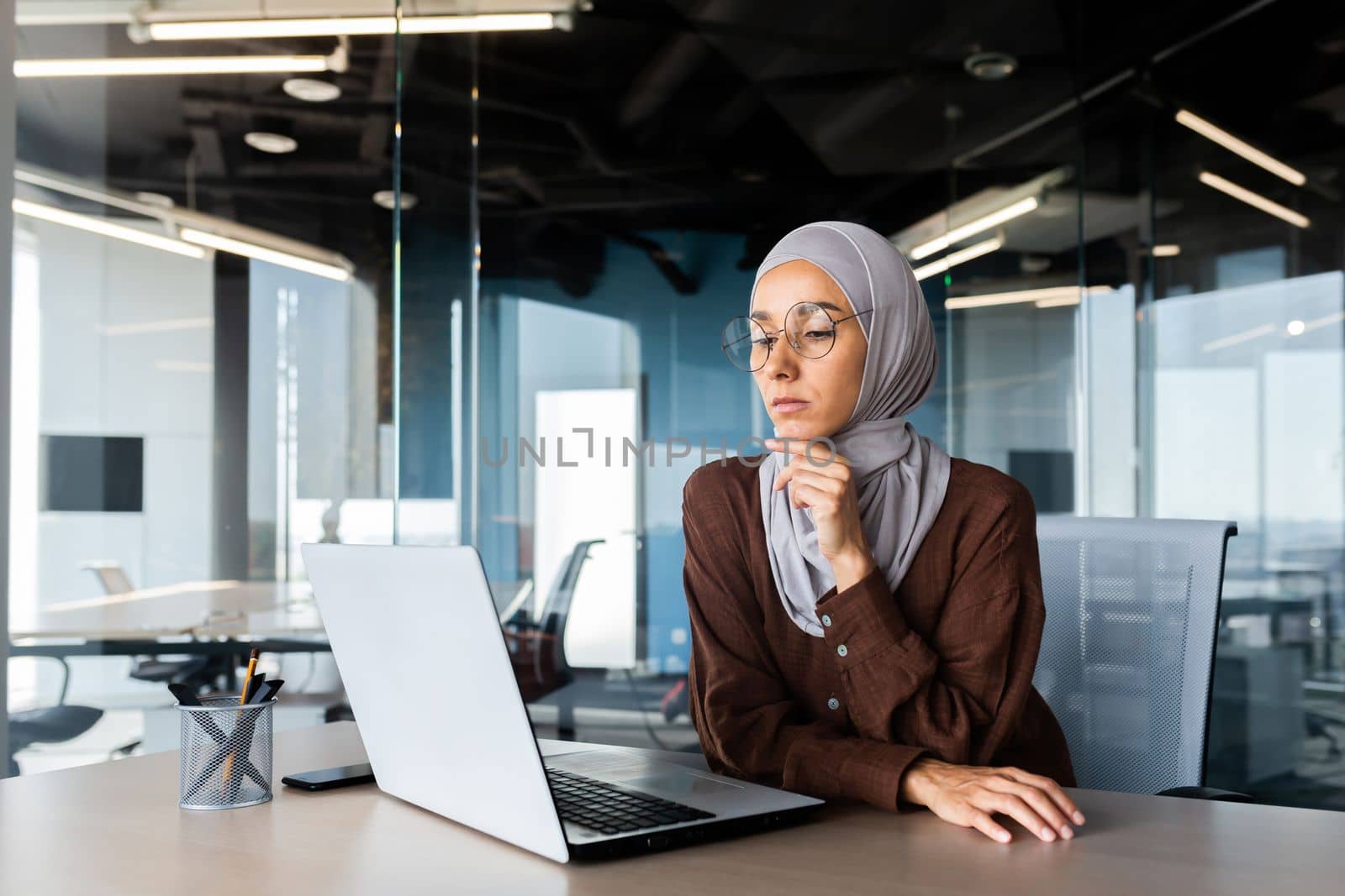 Serious bored businesswoman inside office, muslim woman in hijab thinking while sitting at workplace with laptop, woman at work thinking about decisions by voronaman