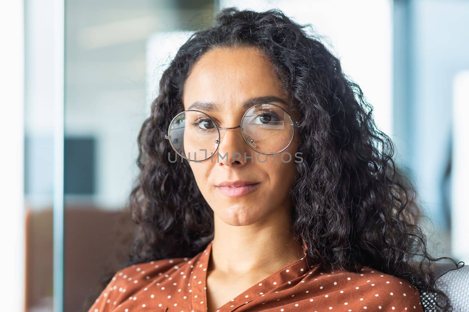 Close-up portrait of confident serious business woman, hispanic woman wearing glasses inside office looking at camera with curly hair.