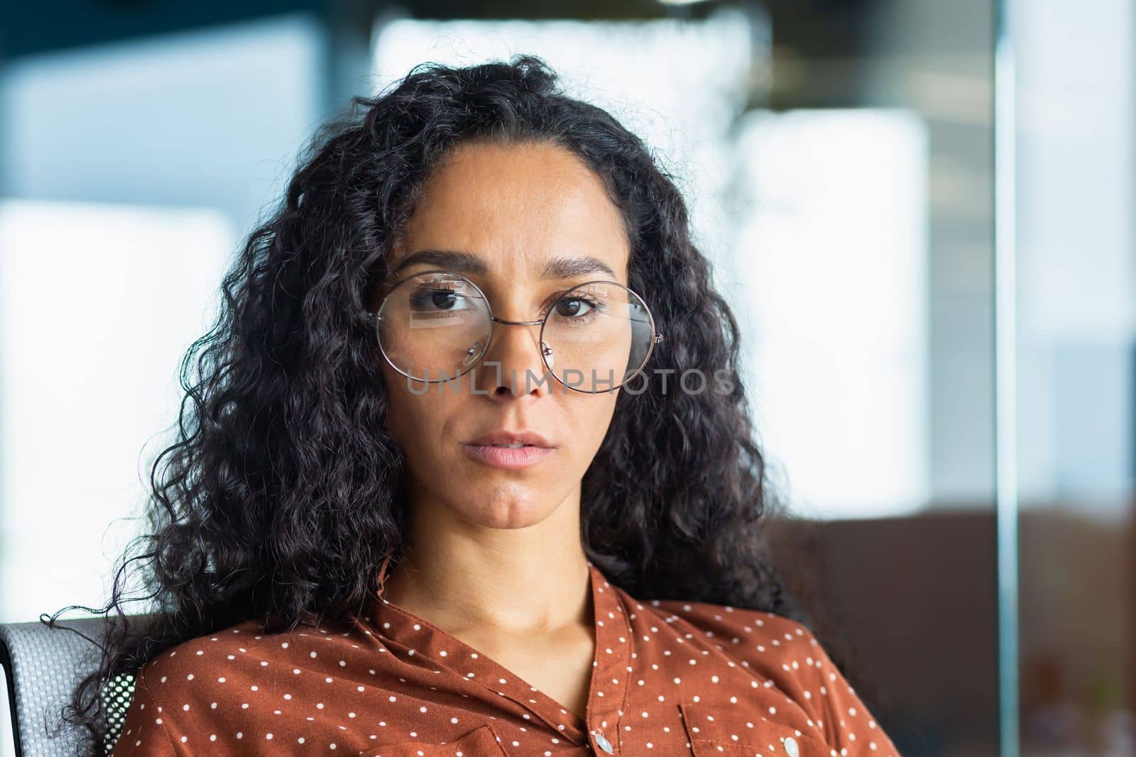 Close-up portrait of confident serious business woman, hispanic woman wearing glasses inside office looking at camera with curly hair.