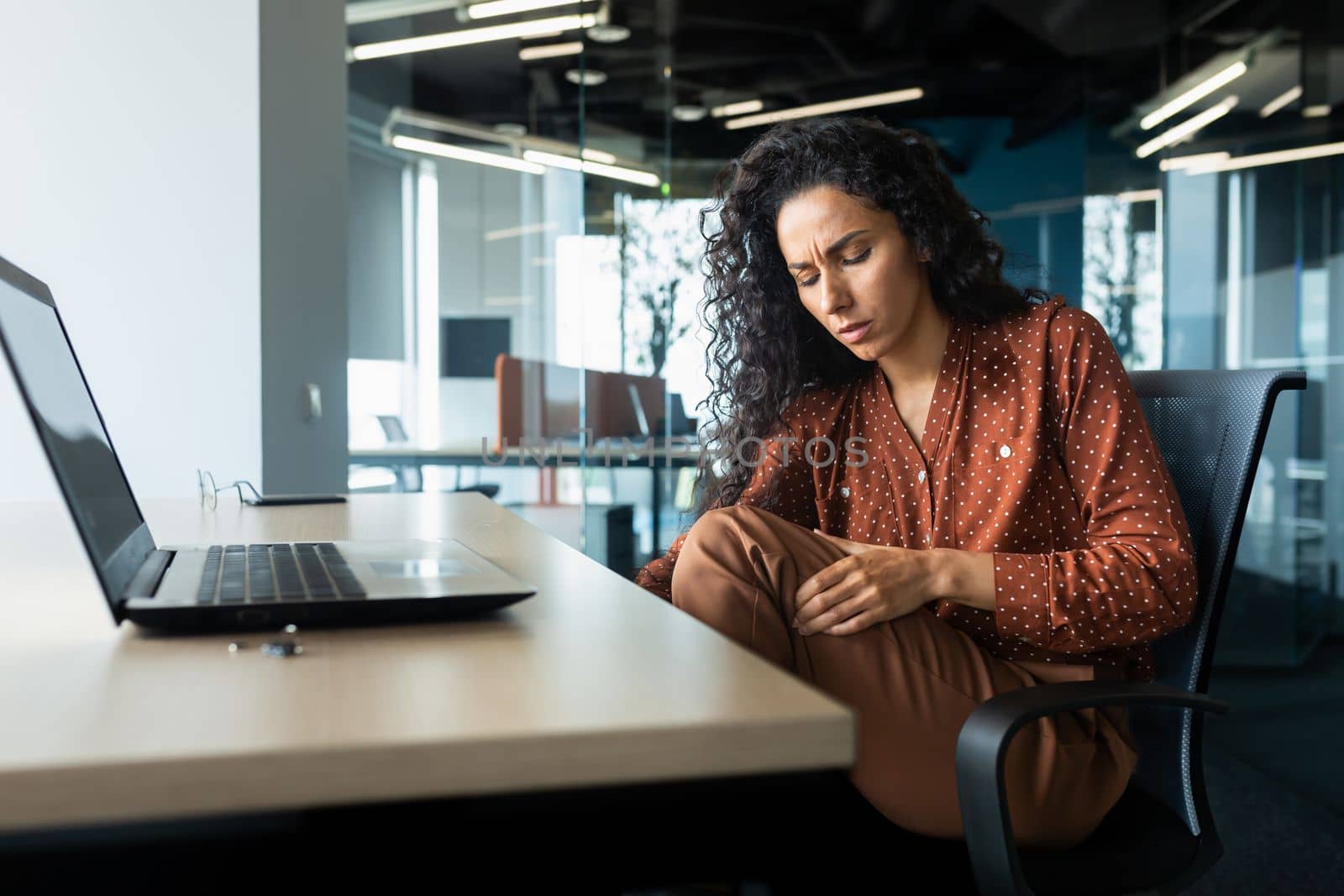 Latin American woman working inside office, business woman has severe leg pain, massaging muscle while sitting at table on chair, using laptop at work.