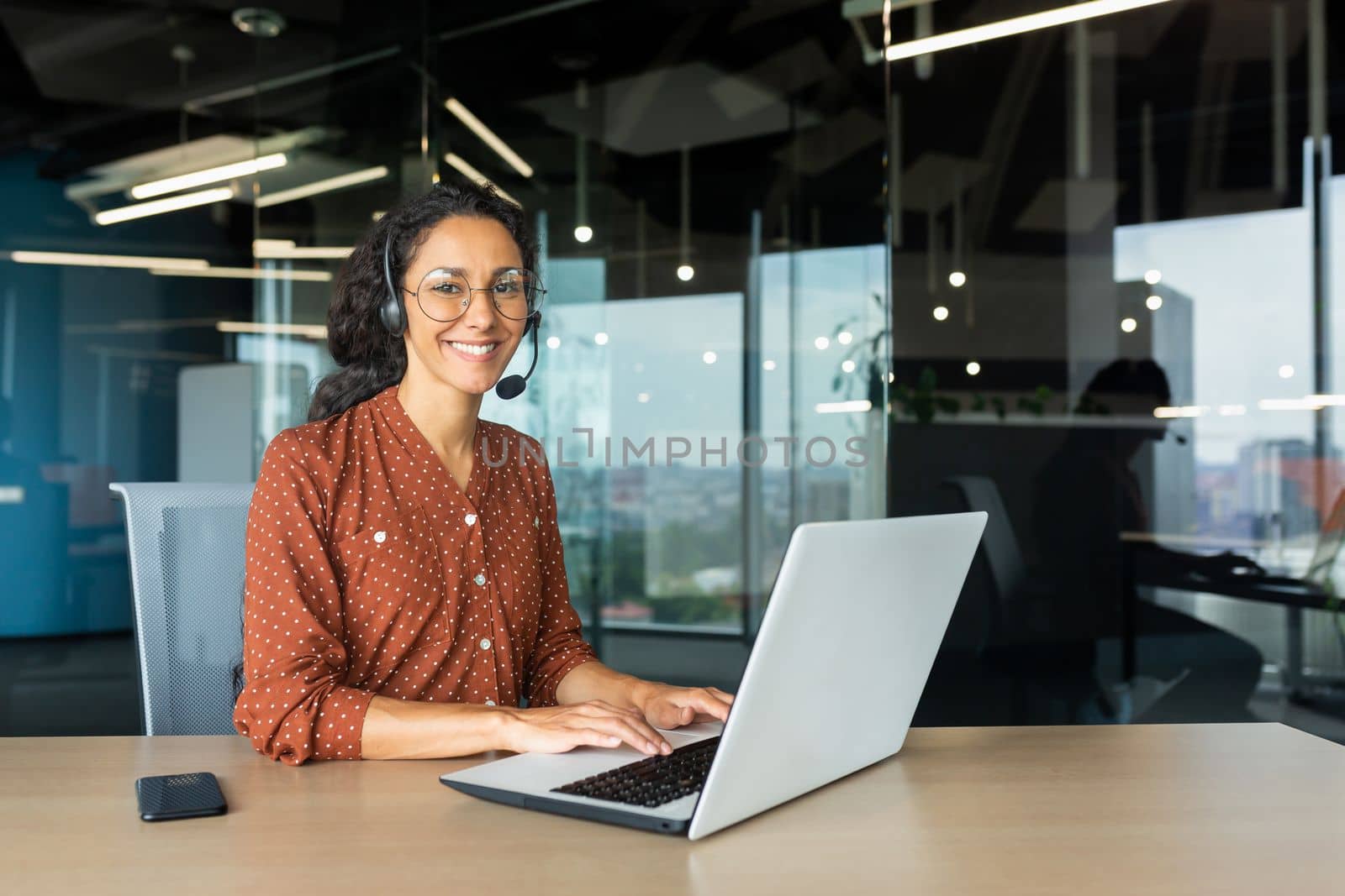 Portrait of Latin American business woman, office worker looking at camera and smiling, using headset and laptop for remote online communication, customer support tech call center worker.