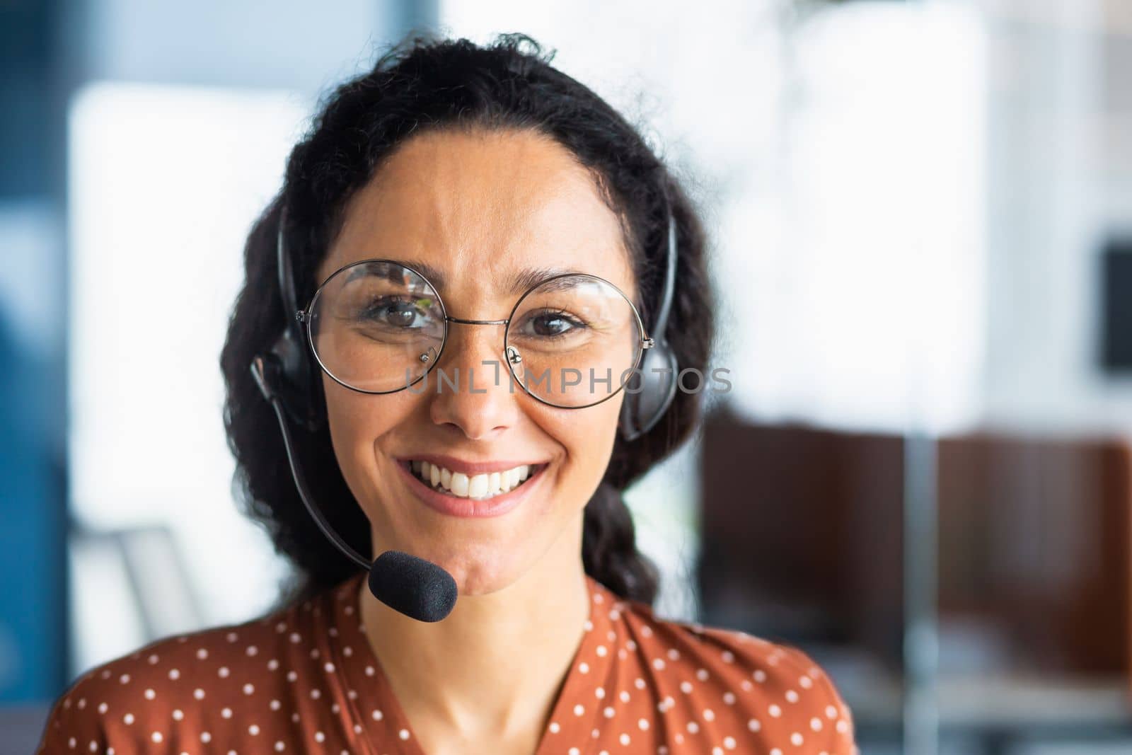 Close up portrait of latin american woman inside modern office with headset for video call, woman smiling and looking at camera, office worker customer support tech helpline.
