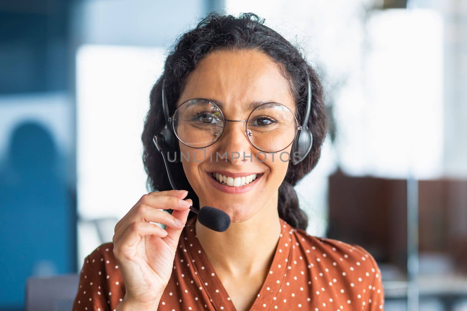 Close up portrait of latin american woman inside modern office with headset for video call, woman smiling and looking at camera, office worker customer support tech helpline.