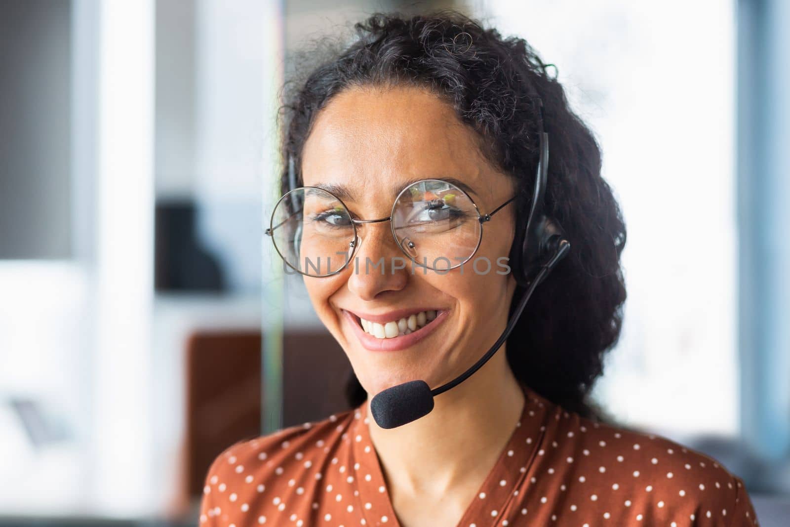 Close up portrait of latin american woman inside modern office with headset for video call, woman smiling and looking at camera, office worker customer support tech helpline.