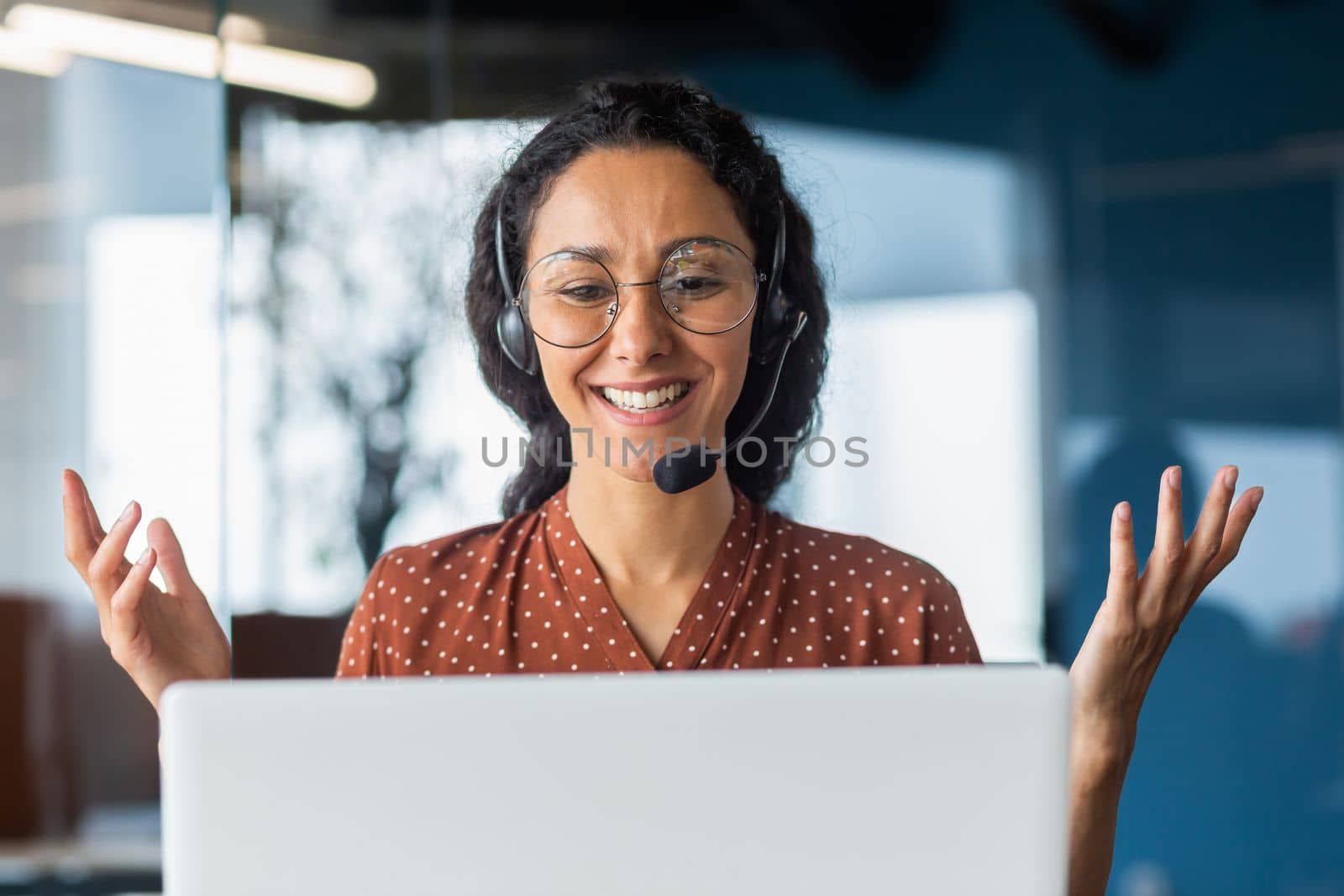 Video call online meeting with colleagues, Hispanic woman working inside modern office, businesswoman smiling and talking remotely using laptop and headset.