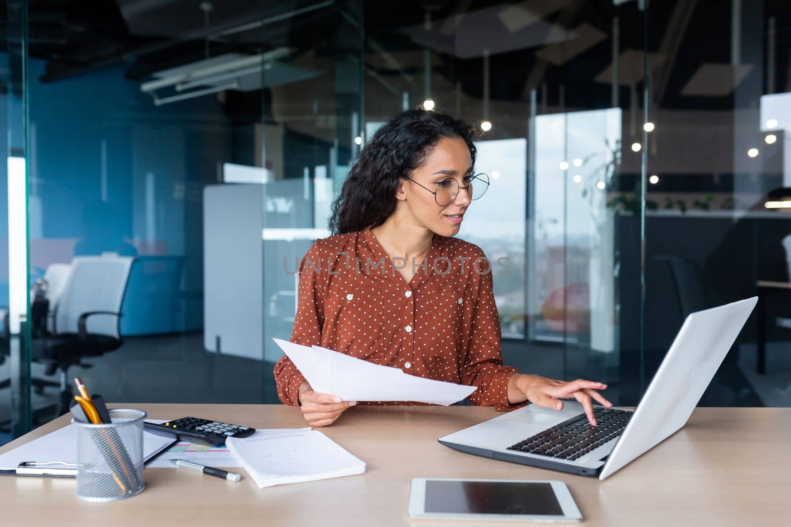 Young happy and successful businesswoman in glasses working with documents inside office, Hispanic woman with laptop looking at bills and contracts, financier with curly hair using laptop.