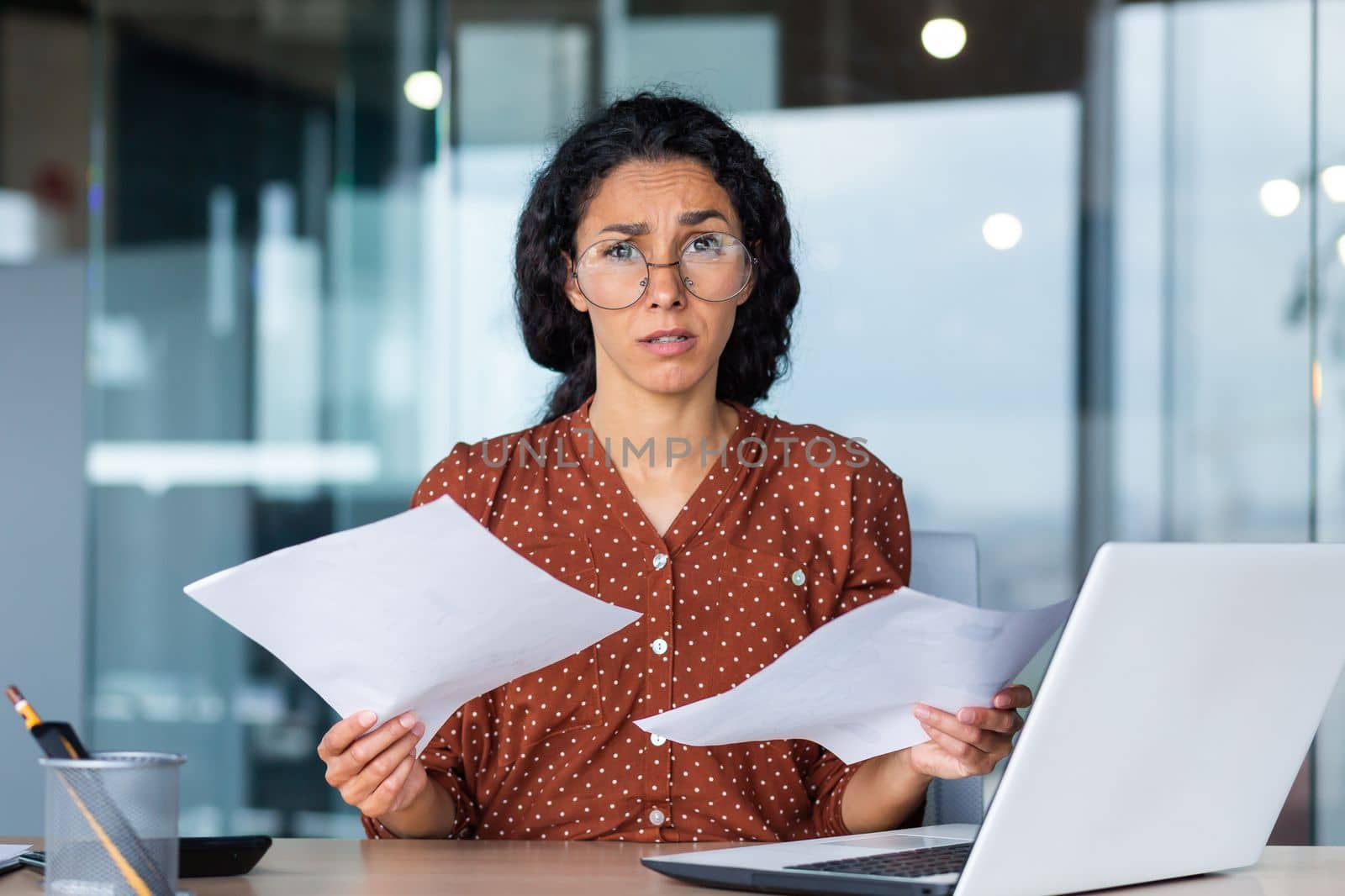 Sad and disappointed woman with documents looking at camera, portrait of upset latin american woman at work, inside office with laptop and paperwork.