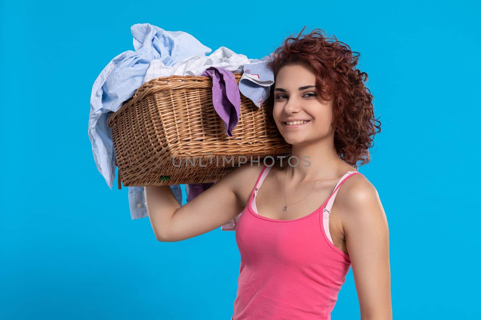 Smiling woman holds on her shoulder a wicker basket containing shirts for washing. Dark curly hair. Daily housework.