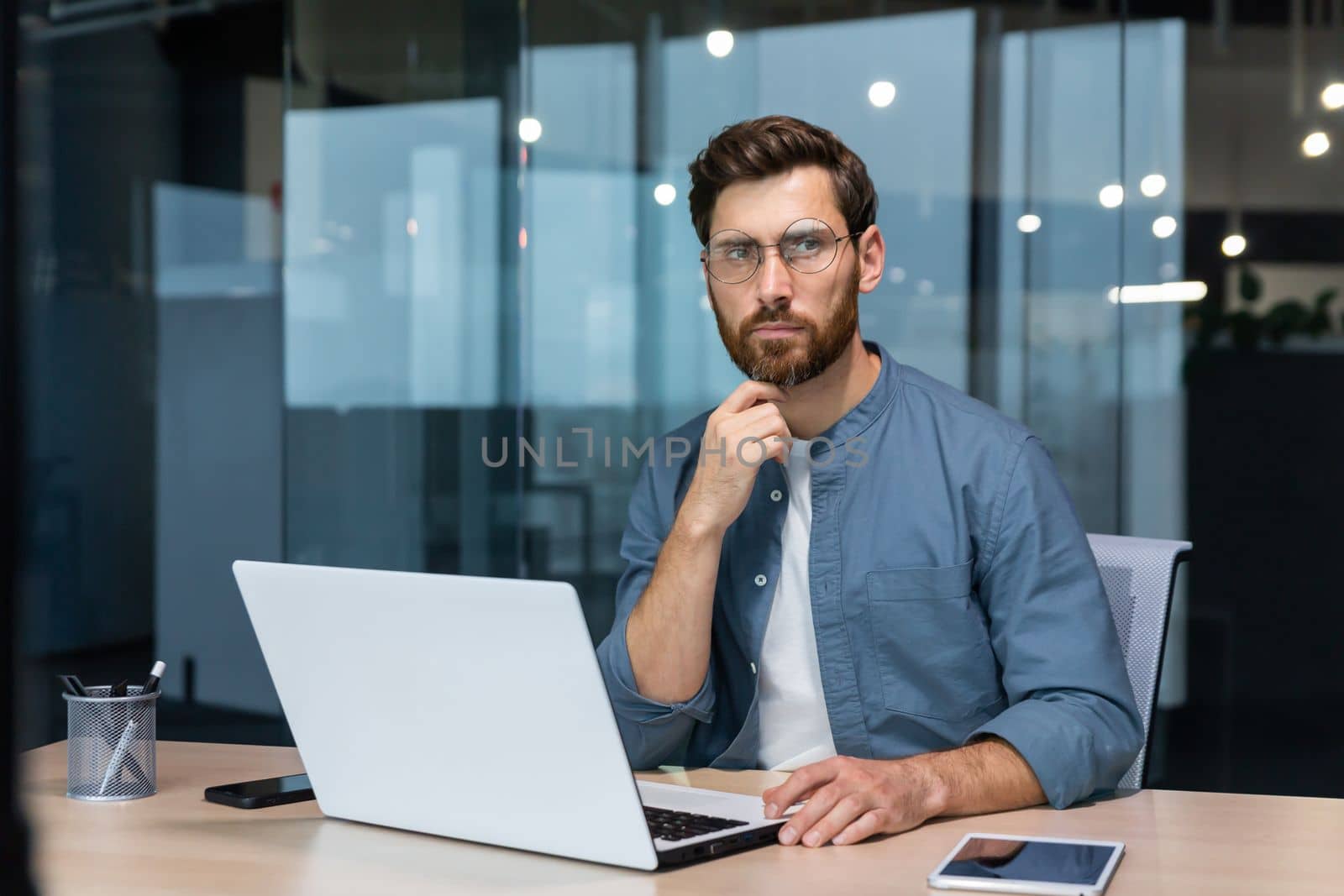 .Serious pensive businessman in shirt thinking about decision sitting at table in modern office, man with beard is using laptop at work.