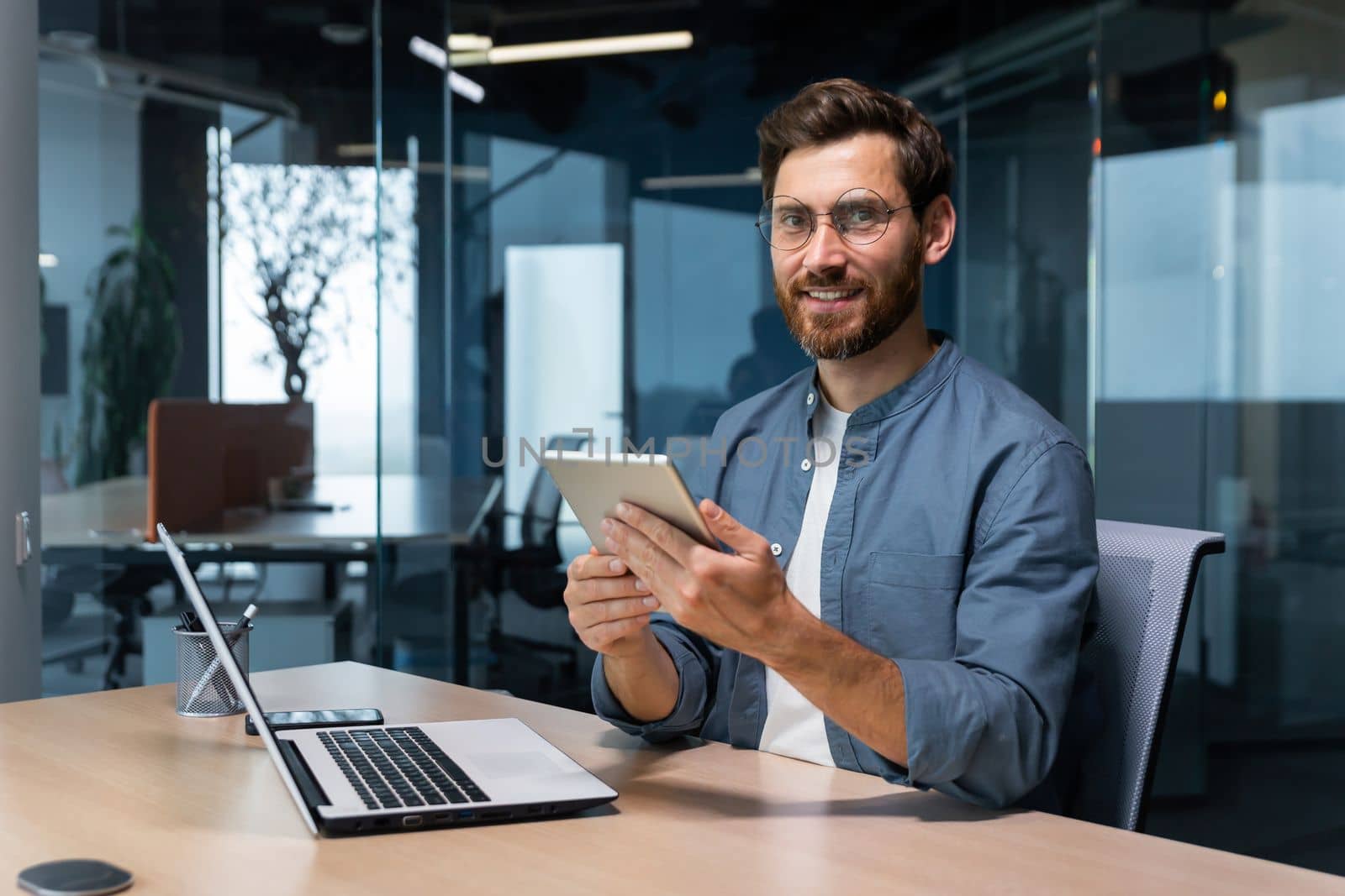 Portrait of a businessman in a modern office, a man uses a tablet computer, sits at a desk, smiles and looks at the camera in a casual shirt and glasses.