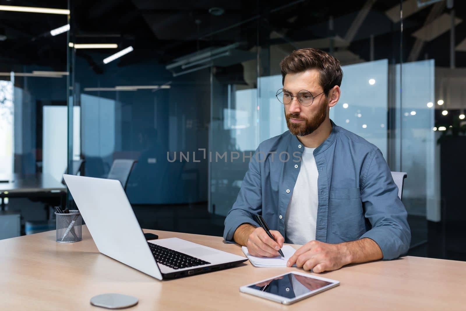 Serious businessman is writing down information studying online while sitting inside the office.